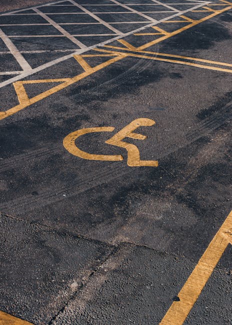 A close-up view of a parking lot surface showing yellow painted wheelchair symbols and parking space markings on dark asphalt. The markings include a large wheelchair icon in the foreground and additi